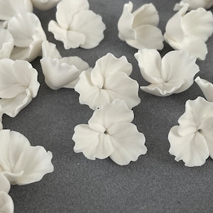 Könnte beinhalten: Eine Ansammlung weißer Keramik-Hortensienblütenköpfe. Die Blütenblätter sind mit feinen Adern versehen, und die Blüten sind auf einer grauen Oberfläche angeordnet. Diese zarten floralen Elemente eignen sich ideal für Bastel- und Dekorationsprojekte.