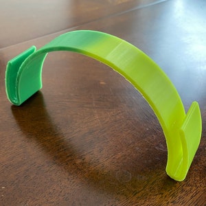 May include: A green and yellow plastic headband with a curved design. The headband is made of a flexible material and has a smooth finish.