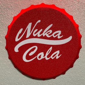 May include: A red bottle cap with the words "Nuka Cola" in white script. The cap has a textured surface and a scalloped edge. The image is a close-up shot.