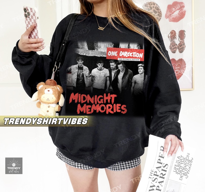 A person wearing a black sweatshirt with a graphic of the One Direction album cover for "Midnight Memories" and holding a stuffed animal and a newspaper.