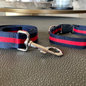 May include: A navy blue pet leash and collar set featuring a bold red horizontal stripe running through the center. The leash includes a silver-toned metal swivel snap hook, while the collar is equipped with a black plastic side-release buckle.