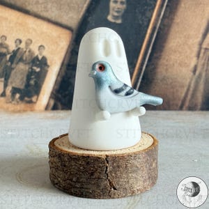 May include: A decorative ceramic ghost figure with a blue pigeon perched on it, set on a wooden base. The pigeon has red eyes and black markings. The ghost is white. The wooden base is a cross-section of a tree trunk. The design is whimsical.