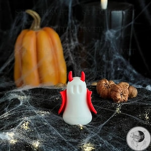 May include: A Halloween-themed still life featuring a white devil-shaped figurine with red horns and wings. An orange pumpkin and several small brown gourds are in the background, all set against a backdrop of faux spiderwebs.