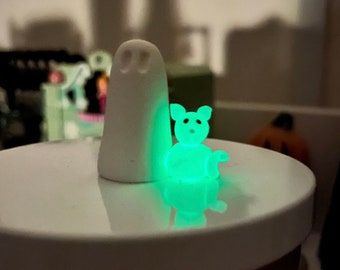 May include: A white ghost figurine with a green glow-in-the-dark cat figurine beside it. Both figurines are on a white surface.