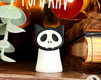 May include: A black and white cat-shaped figurine with a skull face design. The figurine has a white body and a black head with cat ears. The face features large oval eye sockets and a small mouth with two fangs. The figurine is on a wooden surface.