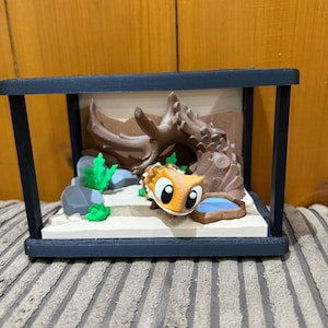 May include: A miniature terrarium featuring a cartoon lizard in orange and white. The scene includes a sandy base, rocks, green plants, and a brown log, all within a black frame. The lizard has large, expressive eyes.