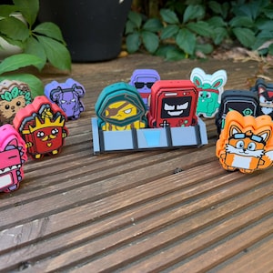 May include: A collection of colourful, small, character-shaped silicone charms featuring various cartoon-style faces and designs. These items include figures in shades of pink, red, orange, blue, and black, some displayed in a grey plastic stand.