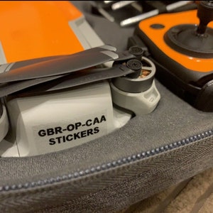 May include: A close-up of a drone with orange and grey accents. The drone is in a grey carrying case. A sticker on the drone reads "GBR-OP-CAA STICKERS".
