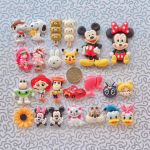 May include: A selection of colourful resin character embellishments, featuring Mickey and Minnie Mouse, Snoopy, and other cartoon figures. The figures are arranged on a light blue patterned surface.