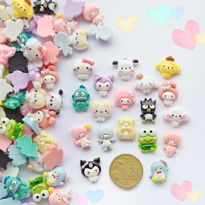 Könnte beinhalten: Eine Sammlung kleiner, bunter Sanrio-Charakter-Cabochons aus Kunstharz in verschiedenen Formen und Pastelltönen, darunter Hello Kitty, Kuromi und Keroppi, die neben einer goldenen Zwei-Dollar-Münze als Größenvergleich auf weißem Grund liegen.