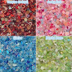 May include: A collection of four distinct assortments of small decorative craft embellishments. The top left quadrant labelled Vampire Cocktail features red, black, and gold pieces. The top right labelled Smoky Pink contains various pink and silver floral shapes. The bottom left labelled Glacier displays blue and clear translucent items. The bottom right labelled Matcha Latte includes green and white floral and leaf-shaped charms.