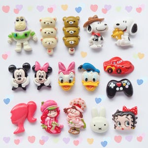 Puede incluir: Una colección de cabujones planos de resina de colores con varios personajes de dibujos animados, incluyendo Buzz Lightyear, Snoopy, Mickey y Minnie Mouse, Donald y Daisy Duck, Lightning McQueen, un mando de juego y Tarta de Fresa.