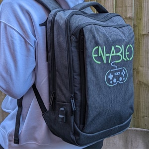 May include: A grey backpack with black accents and a green and white graphic of a video game controller with the text "EN-ABLED".