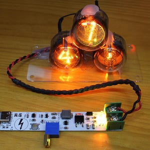 May include: A close-up of an electronic device with three glowing vacuum tubes displaying the number 7, 4, and a blank space. A small screen displays voltage and current readings. The device is connected to a circuit board with a lightning bolt symbol.