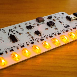 May include: A white Decade Chaser HV555 Nixie Tube Driver circuit board with various electronic components and a USB cable. The board has a series of illuminated orange lights and the text "2026" and "Decade Chaser HV555 Nixie Tuber Driver".