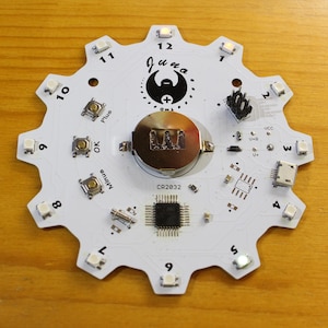 May include: A white circuit board with a circular design featuring a black logo with the text "Juna" and a plus symbol. The board has 12 white LEDs, three buttons labeled "Minus", "OK", and "Plus", and a battery compartment labeled "CR2032".