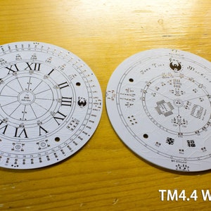May include: Two white circular circuit boards with intricate designs. One features Roman numerals and a clock face, the other has text and symbols. The boards have numerous small holes and electronic components. The text "TM4.4 White" is visible.