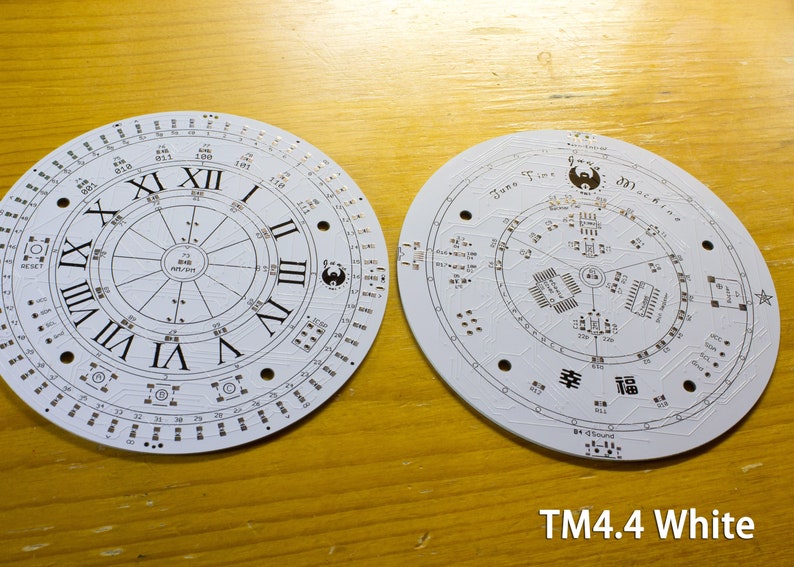 May include: Two white circular circuit boards with intricate designs. One features Roman numerals and a clock face, the other has text and symbols. The boards have numerous small holes and electronic components. The text "TM4.4 White" is visible.