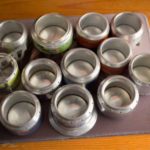 May include: A collection of 12 silver aluminum cans with the tops and bottoms removed, arranged on a gray surface. The cans are used for making DIY candle holders.