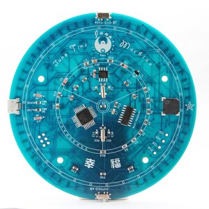 May include: A circular teal circuit board with the text "June Time Machine" and various electronic components. The board features intricate circuitry patterns, with the word "幸福" and other text. The board is approximately 5 inches in diameter.