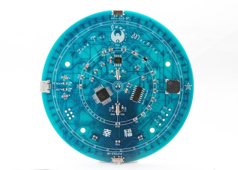 May include: A circular teal circuit board with the text "June Time Machine" and various electronic components. The board features intricate circuitry patterns, with the word "幸福" and other text. The board is approximately 5 inches in diameter.