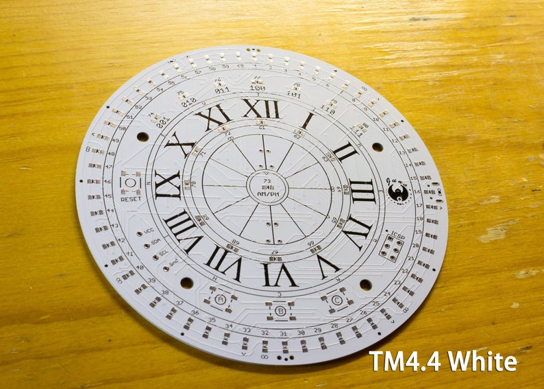 May include: A white circular electronic circuit board with Roman numerals marking the hours. The board has numerous small components and the text "TM4.4 White". The design includes the text "RESET" and other technical markings.