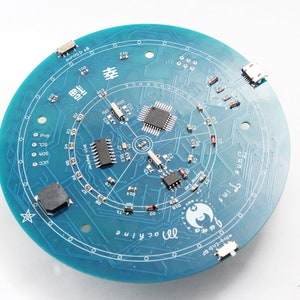 May include: A circular teal circuit board with various electronic components, including chips, resistors, and connectors. The board features intricate circuit patterns and text, including "June", "machine", and "Pro-CAD".
