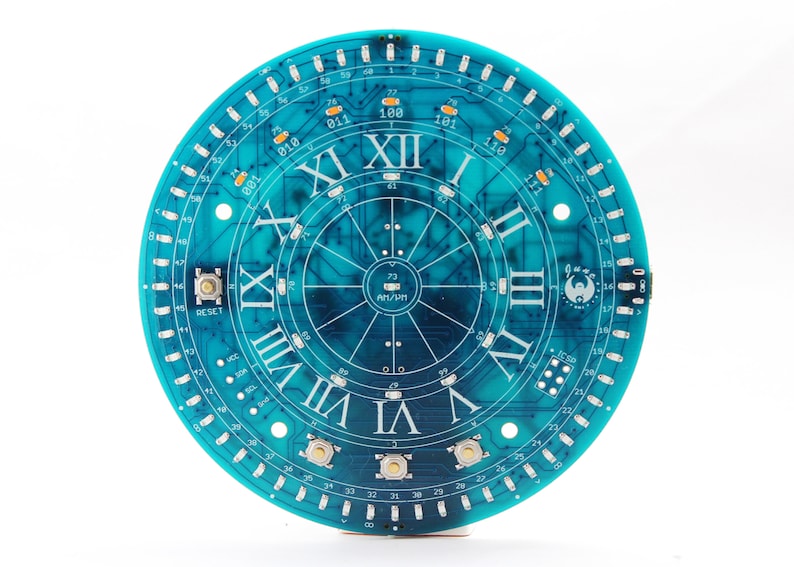 May include: A teal-colored circular circuit board with Roman numerals marking the hours. The board features electronic components, buttons, and the text "RESET" and "AM/PM". The design resembles a clock face.