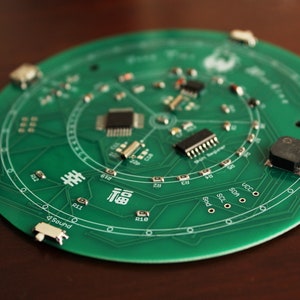 May include: A circular green circuit board with various electronic components, including chips and resistors. The board has white text and symbols, including the word "Sound" and Chinese characters. The board is on a brown surface.
