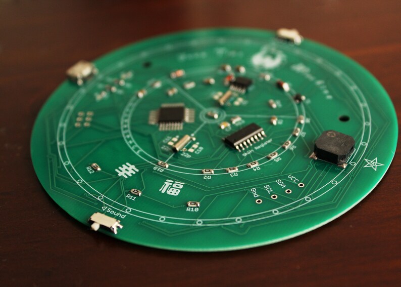 May include: A circular green circuit board with various electronic components, including chips and resistors. The board has white text and symbols, including the word "Sound" and Chinese characters. The board is on a brown surface.