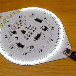 May include: A white circuit board with a circular design and the text "Juno Century Chaser 2026". The board has various electronic components, a crescent moon symbol, and a USB cable connected to it.