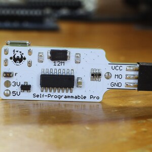 May include: A white circuit board with black components and the text "Self-Programmable Pro". The board has a micro USB connector, a 12M resistor, and a 512 resistor. The board has a black header connector on the right side.