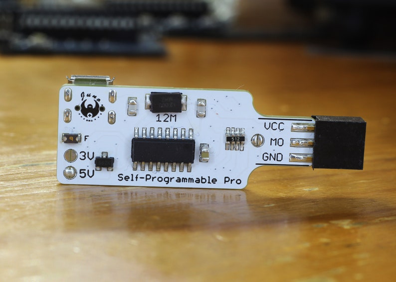 May include: A white circuit board with black components and the text "Self-Programmable Pro". The board has a micro USB connector, a 12M resistor, and a 512 resistor. The board has a black header connector on the right side.