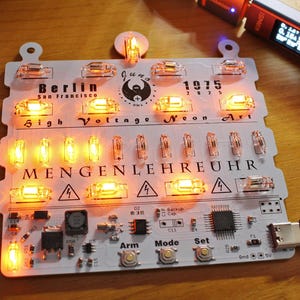 May include: A lit-up circuit board with the text "Berlin San Francisco 1975 High Voltage Neon Art" and "MENGENLEHREUHR". The board has several glowing orange lights and is connected to a USB cable.