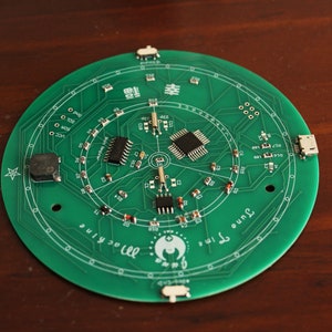 May include: A circular green circuit board with various electronic components, including chips, resistors, and connectors. The board has a printed design with the text "June Time Machine" and Chinese characters. The board is on a brown surface.