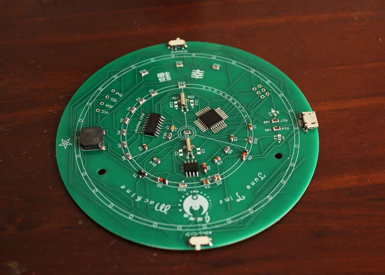 May include: A circular green circuit board with various electronic components, including chips, resistors, and connectors. The board has a printed design with the text "June Time Machine" and Chinese characters. The board is on a brown surface.