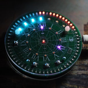May include: A circular green circuit board with Roman numerals and LED lights. The board has a clock face design with a USB cable attached. The LEDs are various colors, including blue, red, and purple.