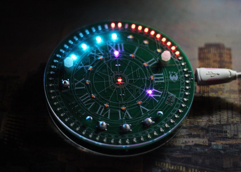 May include: A circular green circuit board with Roman numerals and LED lights. The board has a clock face design with a USB cable attached. The LEDs are various colors, including blue, red, and purple.