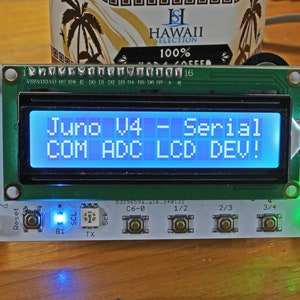 May include: A green circuit board with a blue LCD screen displaying the text "Juno V4 - Serial COM ADC LCD DEV!" The board has a green power LED lit, a blue LED lit, and a reset button. There are also several buttons labeled "C6-0", "1/2", "2/3", and "3/4".