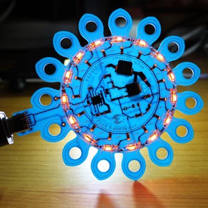 May include: A blue electronic circuit board with a circular design and a series of orange lights. The board is connected to a device displaying voltage and current readings. The design includes the text "Precision Micro" and "June Sunflowers".