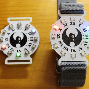 May include: Two white electronic watches with Roman numeral hour markers and a black emblem. One watch is on a gray strap. Both watches have colored LED lights and are on a wooden surface.
