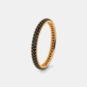 Peut inclure: Une bague dorée entièrement sertie de petites pierres noires. La bague est présentée sur un fond clair et uni. L'intérieur de la bague est également visible, mettant en valeur le savoir-faire et le design.