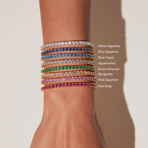Può includere: Una collezione di braccialetti dorati, ognuno ornato da una gemma di colore diverso. Le gemme includono zaffiro bianco, zaffiro blu, topazio blu, acquamarina, smeraldo verde, morganite, zaffiro rosa e rubino rosso. I braccialetti sono impilati su un polso.