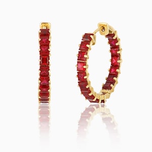 Puede incluir: Un par de pendientes de aro dorados, cada uno incrustado con vibrantes piedras preciosas rojas de corte cuadrado. Los pendientes se exhiben sobre una superficie reflectante, mostrando su diseño y color.