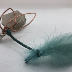 May include: A copper wire hair clip with a crystal attached and a teal feather attached to the end of the clip.