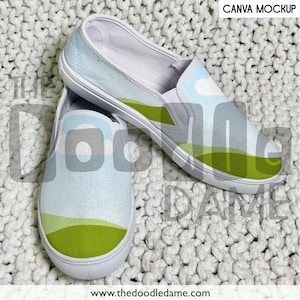 May include: A pair of white canvas slip-on shoes with a green, blue, and white landscape design. The shoes are on a white fuzzy surface.