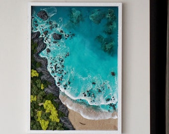 May include: A framed resin art piece depicting an aerial view of a beach with turquoise water, white waves, and green foliage. The beach is surrounded by dark gray rocks.