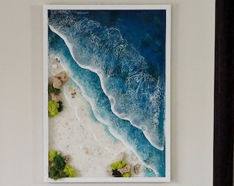 Puede incluir: Una pieza de arte de resina enmarcada que representa una escena de playa con olas azules del océano rompiendo en una costa de arena blanca. Las olas están superpuestas y texturizadas, creando un efecto 3D. Musgo verde y pequeñas conchas blancas están esparcidas por la arena.
