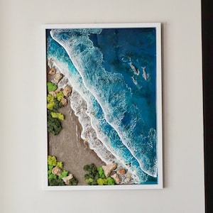 May include: A framed resin art piece depicting an aerial view of a beach with turquoise water, white waves, and green moss. The beach is bordered by a rocky shore with green moss and small rocks.