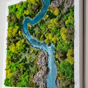 Puede incluir: Una pieza de arte de musgo enmarcada que representa un r&iacute;o serpenteando a trav&eacute;s de un exuberante bosque verde. El r&iacute;o es de un azul vibrante y el musgo es de varios tonos de verde.